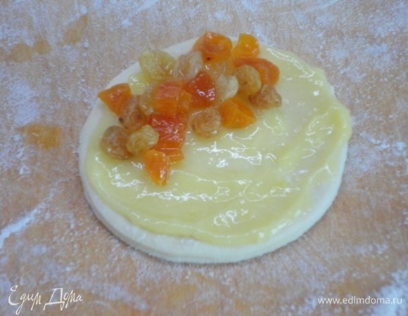 Смажьте лепешечки заварным кремом. Часть крема у вас останется — с ним будете есть готовый пирог. На половину каждого кружочка насыпьте по ложечке изюма.