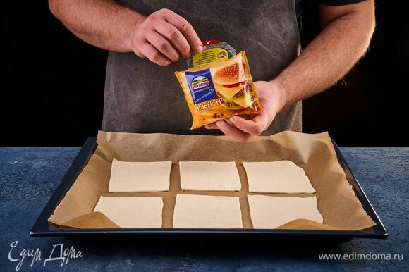 Разрежьте тесто на 6 квадратов. На противень, застеленный пергаментом, выложите заготовки. Возьмите плавленый сыр Hochland Чизбургер.