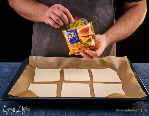 Разрежьте тесто на 6 квадратов. На противень, застеленный пергаментом, выложите заготовки. Возьмите плавленый сыр Hochland Чизбургер.