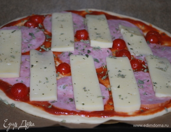 Тонко раскатайте тесто на пергаменте для выпечки. Ветчину и сыр нарежьте на тонкие ломтики. Тесто смажьте томатным соусом, посыпьте сушеным базиликом, сверху разложите ветчину, сыр и помидоры. Выпекайте в предварительно разогретой духовке при 180°C примерно 15–20 минут.