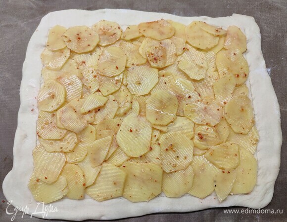 Чеснок натереть на мелкой терке и распределить его по тесту. Картофель нарезать овощечисткой на слайсы. Посыпать солью и любимыми специями. Разложить слайсы на тесто.