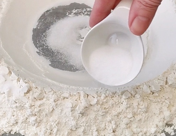 Высыпаем просеянную муку на рабочую поверхность. Делаем в центре углубление. В него отправим сахар, а соль распределим равномерно по краям муки.