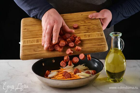 На сковороде с растительным маслом обжарьте лук и морковь. Добавьте нарезанные колбаски, потушите.