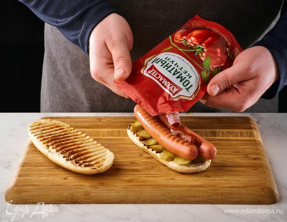 На булочки выложите кружочки огурцов, сосиски, полейте томатным кетчупом «Кухмастер».