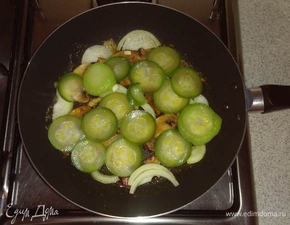 Далее к чесноку и грибам добавить репчатый лук и кабачок. Перемешать все. Убрать огонь до минимума и обжаривать овощи до готовности, помешивая.