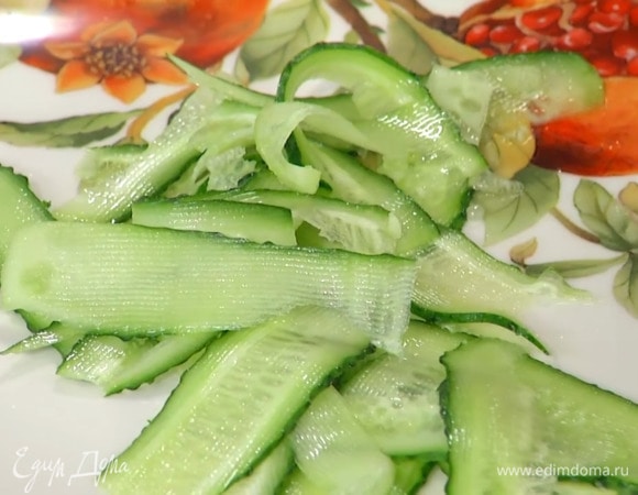 Огурец с помощью овощечистки нарезать тонкими полосками и выложить в большую тарелку, посыпать измельченным луком и листьями тархуна.