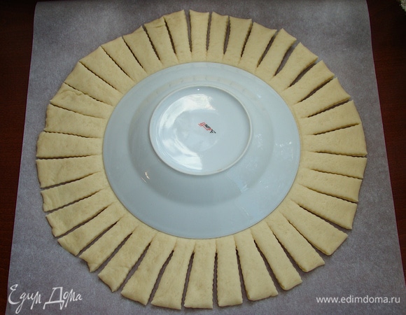 Тесто раскатайте в большой круг (лучше это сделать сразу на бумаге для выпечки). Диаметр моей ватрушки — 32 см, толщина — 0,7 см. В середину уложите крышку, тарелку или любой круглый предмет и сделайте по всему кругу надрезы шириной 1,5–2 см.