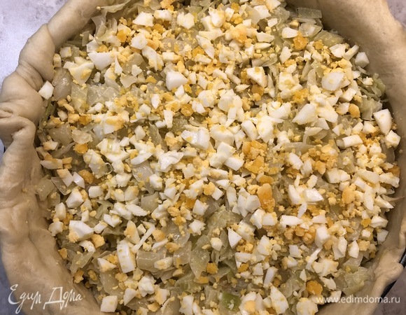 Форму для выпечки смазать сливочным маслом. Готовое тесто разделить на 2 части: большую и меньшую. Большая часть — дно и стенки пирога, меньшая — верх пирога. Из большей части теста раскатать круг и выложить его в форму, чтобы края немного свисали. Внутрь выложить всю капустную начинку, на нее — мелко нарубленные яйца.