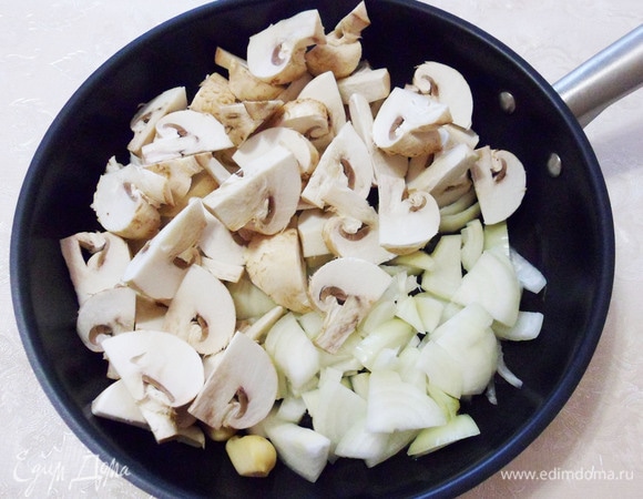 На разогретую сковороду выложите репчатый лук, нарезанный полукольцами, шампиньоны и зубчики чеснока.