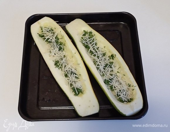 Достать противень с кабачками и выложить на дно каждой половинки измельченные чеснок и укроп. Затем посыпать каждую половинку кабачка половиной тертого сыра.