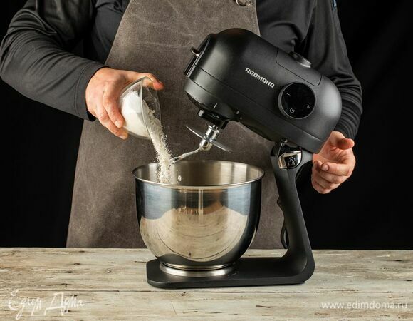 Установите на кухонную машину насадку для замешивания теста. Муку соедините с сухими дрожжами и добавьте в чашу кухонной машины. С помощью насадки крюк замесите пластичное тесто.