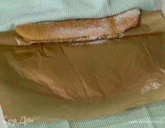 Оставшимся сахаром присыпать кухонное полотенце по всему периметру, перевернуть на него теплый выпеченный корж и отделить пекарскую бумагу, затем снова накрыть ей корж и с помощью полотенца свернуть все в рулет.