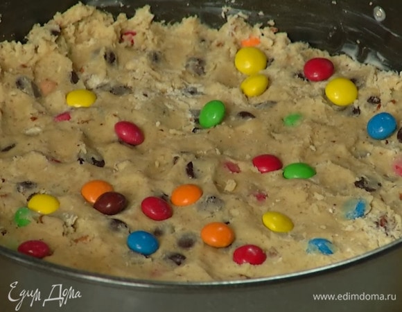 Дно разъемной формы диаметром 28 см выстелить бумагой для выпечки и смазать оставшимся сливочным маслом, затем равномерно выложить тесто, уплотнить его руками, посыпать оставшимся разноцветным драже и слегка утопить его.