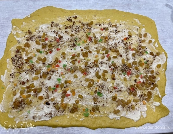 Раскатайте первую половину теста в прямоугольник. Чем он будет тоньше, тем более красивые завитки будут у краффина в итоге. Чтобы тесто опять-таки не липло к рукам и скалке, можно либо немного посыпать поверхность мукой, либо и стол, и скалку смазать растительным маслом. Смажьте тесто половиной сливочного масла, посыпьте сахаром и подготовленной начинкой из орехов и изюма. Цукаты и другие добавки (сушеная вишня, клюква) после выпечки дают свой вкус, поэтому в начинке их можно не использовать или использовать то, что вам нравится.