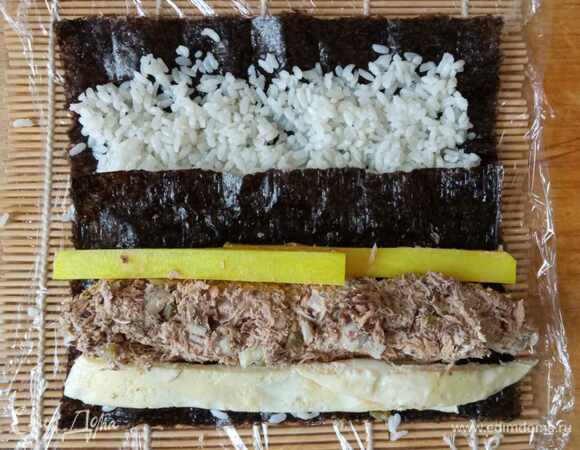 Начинаем собирать кимпаб. Берем лист нори. Кладем на обернутую пищевой пленкой циновку шершавой стороной вверх. Это нужно для лучшего прилипания риса. Лист нори покрываем тонким слоем риса. Рис должен быть чуть теплым, чтобы кимпаб лучше сворачивался. Если у вас рис был приготовлен заранее и уже холодный, можно разогреть его в микроволновке. Снизу оставляем примерно 1 см без риса и так же сверху. Для кимпаба с тунцом нам нужна еще половинка листа нори, которую мы кладем поверх риса в нижней части. Это необходимо, чтобы тунец не размягчил водоросли. Выкладываем сверху тунца, с одной стороны кладем дайкон, с другой — яичную полоску. Желательно, чтобы дайкон был размером с лист нори, но в этот раз я смогла купить только небольшой дайкон, поэтому положила два брусочка внахлест.