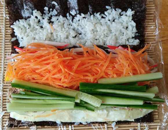 Сверху кладем морковь, крабовые палочки и огурец. С помощью циновки аккуратно, но плотно сворачиваем рулет. Верхний край нори можно чуть-чуть смазать водой, чтобы лучше слиплось.