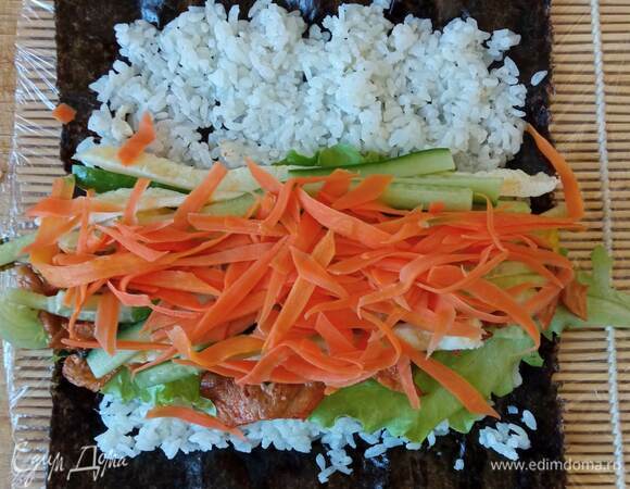 Начинаем собирать кимпаб. Берем лист нори. Кладем на обернутую пищевой пленкой циновку шершавой стороной вверх. Это нужно для лучшего прилипания риса. Лист нори покрываем тонким слоем риса. Рис должен быть чуть теплым, чтобы кимпаб лучше сворачивался. Если у вас рис был приготовлен заранее и уже холодный, можно разогреть его в микроволновке. Снизу оставляем примерно 1 см без риса и так же сверху. На рис кладем внахлест пару листов салата, сверху выкладываем соевое мясо, с одной стороны мяса кладем дайкон, с другой — яичную полоску. Желательно, чтобы дайкон был размером с лист нори, но если короче, берем два брусочка. Сверху кладем морковь и огурец. С помощью циновки аккуратно, но плотно сворачиваем рулет. Верхний край нори можно чуть-чуть смазать водой, чтобы лучше слиплось. Свернутый рулет разглаживаем руками от центра к краям. Смазываем кисточкой кунжутным маслом. Даем полежать пару минут и нарезаем очень острым ножом на кусочки 1–1,5 см шириной. Края кимпаба оставляем как есть с торчащими овощами. При подаче посыпаем кунжутом.