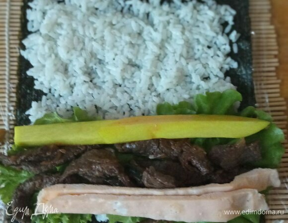 Начинаем собирать кимпаб. Берем лист нори. Кладем на обернутую пищевой пленкой циновку шершавой стороной вверх. Это нужно для лучшего прилипания риса. Лист нори покрываем тонким слоем риса. Рис должен быть теплым, чтобы кимпаб лучше сворачивался. Если у вас рис был приготовлен заранее и уже холодный, можно разогреть его в микроволновке. Снизу оставляем примерно 1 см без риса и так же сверху. На рис выкладываем по 2 листа салата, на него мясо, с одной стороны мяса кладем дайкон, с другой — яичный блинчик.