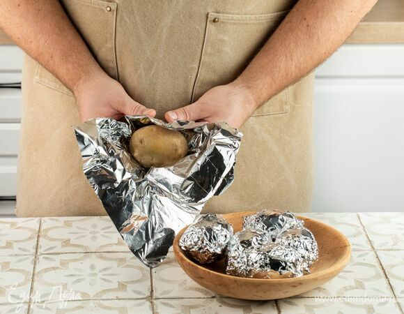 Клубни картофеля хорошо промойте водой и очистите от загрязнений. Заверните в фольгу и проткните вилкой. Отправьте запекаться в духовку на 1 час при температуре 180°С.