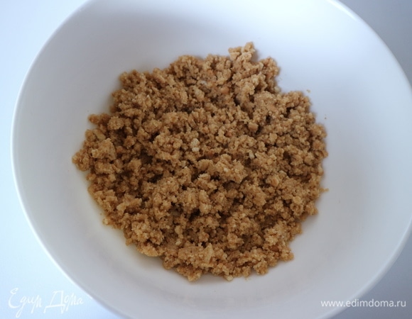 Духовку разогреть до 150°C. Сливочное масло вынуть заранее. Дно формы застелить бумагой для выпечки, бока смазать оливковым маслом. Приготовить основу чизкейка. Измельчить до мелкой крошки печенье, добавить мягкое сливочное масло, перемешать.