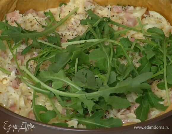 В подпеченный корж выложить половину руколы, залить начинкой, посолить, поперчить и посыпать мускатным орехом, сверху выложить оставшуюся руколу и слегка притопить ее вилкой.