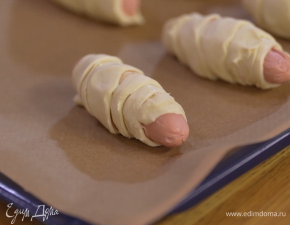 По всей длине полосок теста разложить грудинку, обернуть сосиски тестом по диагонали и выложить на выстеленный бумагой для выпечки противень.