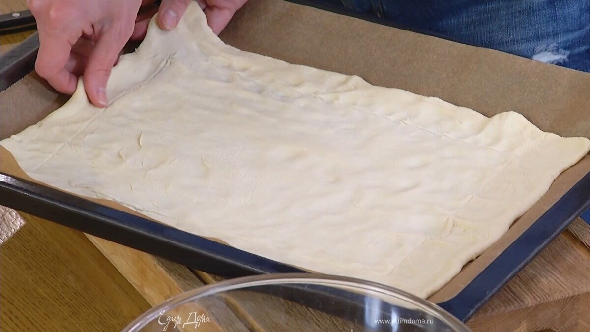 Тесто предварительно разморозить, выложить на выстеленный бумагой для выпечки противень и растянуть руками по всему периметру, затем, не прорезая насквозь и отступив на пару сантиметров от края, сделать ножом надрезы.